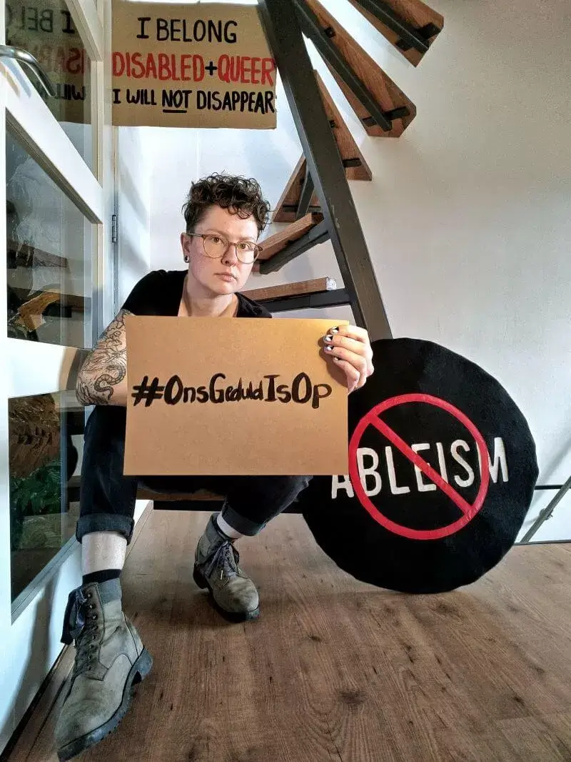 Elina Pollaert houdt een bord vast met #Ons-Geduld-Is-Op. Achter haar een bord met de tekst: I Belong, disabled and queer, I will nog disappear.
