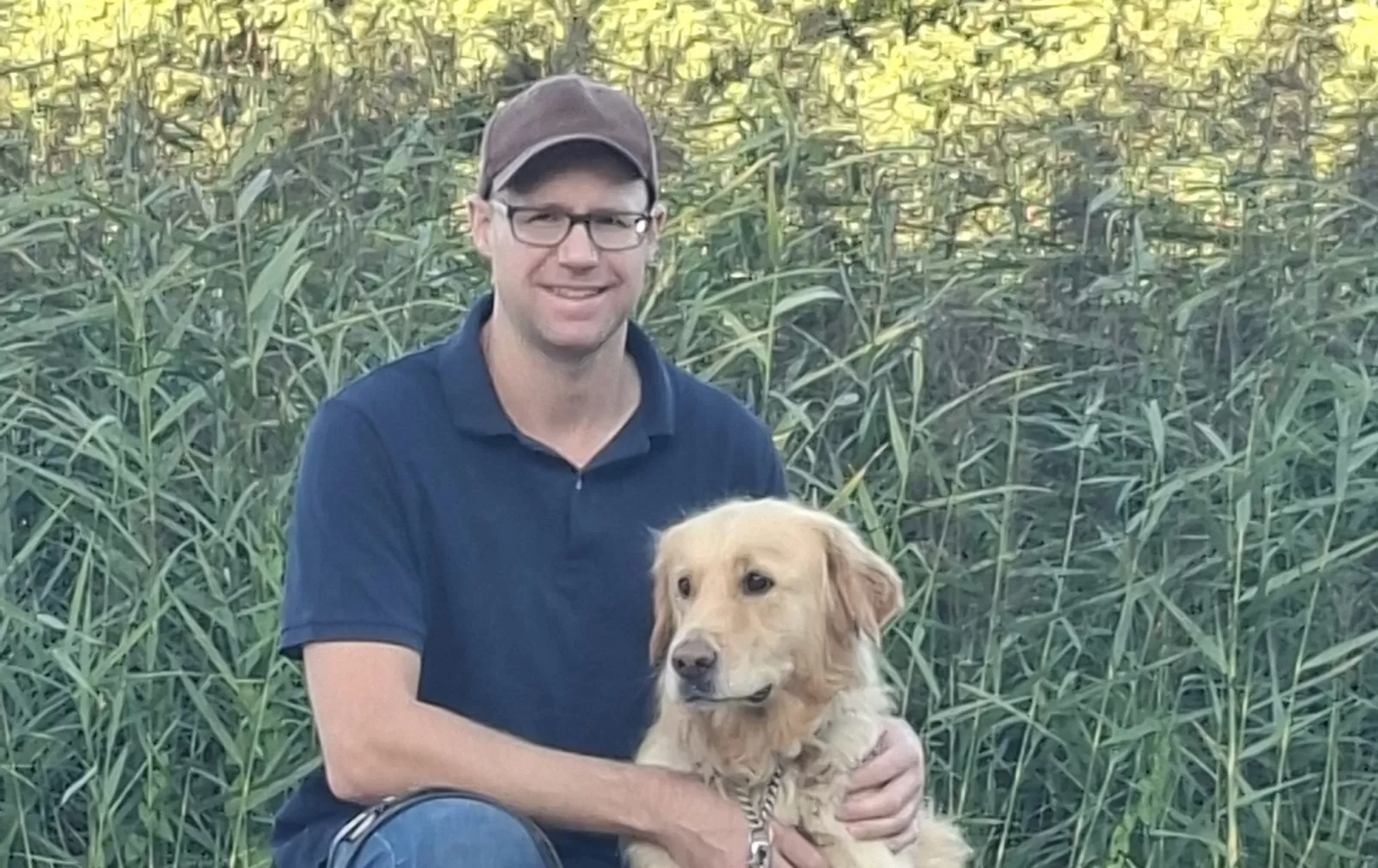 Man in een blauwe polo met een bruine pet en een bril, zit gehurkt naast zijn hond, een golden retriever. Achter hen is riet zichtbaar.