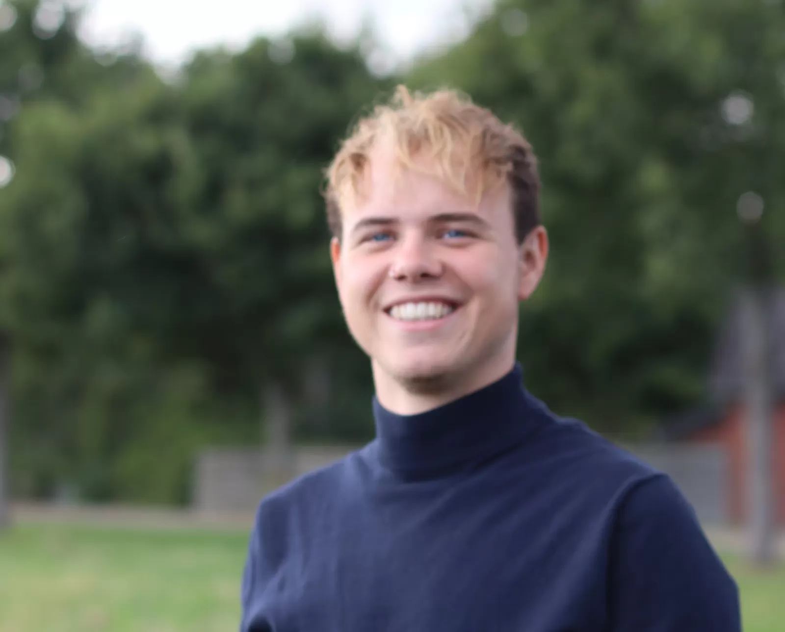 Lachende jongeman met kort blond haar in een zwarte koltrui. Op de achtergrond zijn bomen en gras zichtbaar.