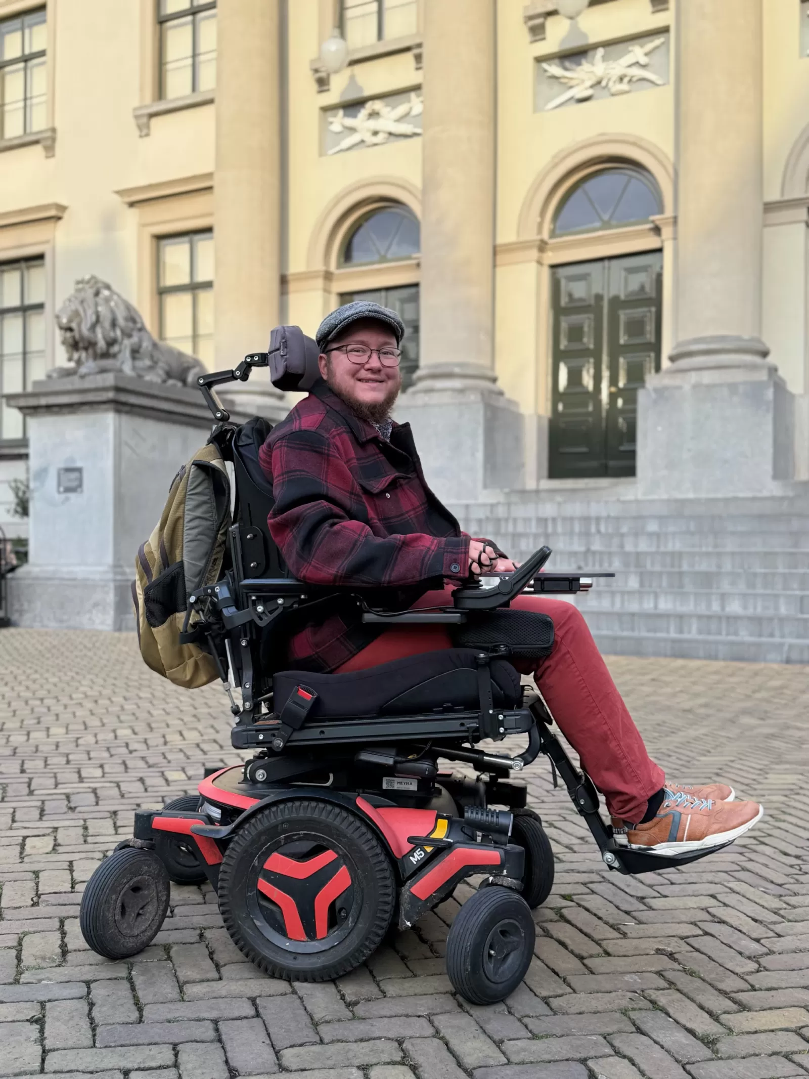 Portret Nick Bootsman. Man in een rolstoel voor het stadhuis in Dordrecht. Hij heeft een baard, draagt een rode broek, rood geruite jas en een grijze cap.