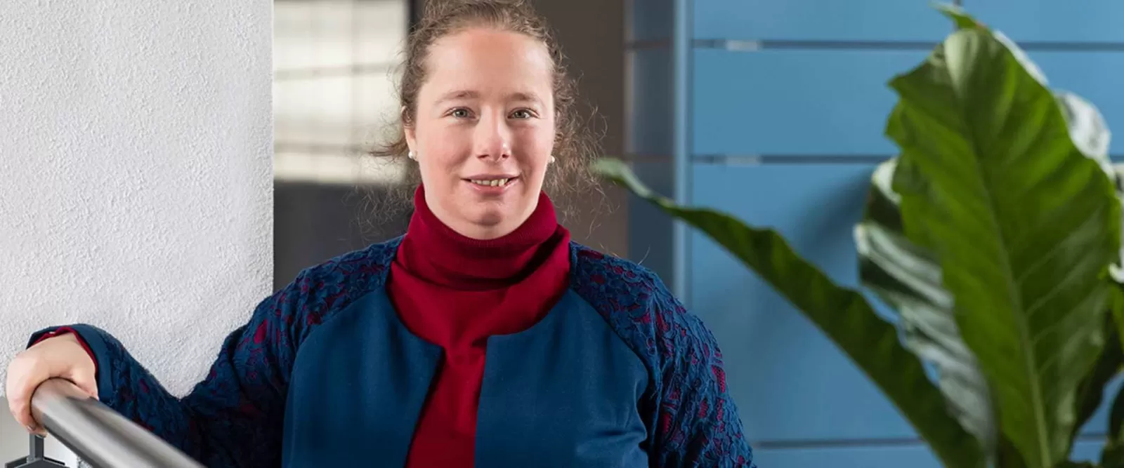 Portret van een vrouw in een blauwe trui en rode sjaal, voor een deels witte, deels blauwe muur en een grote tuinplant.