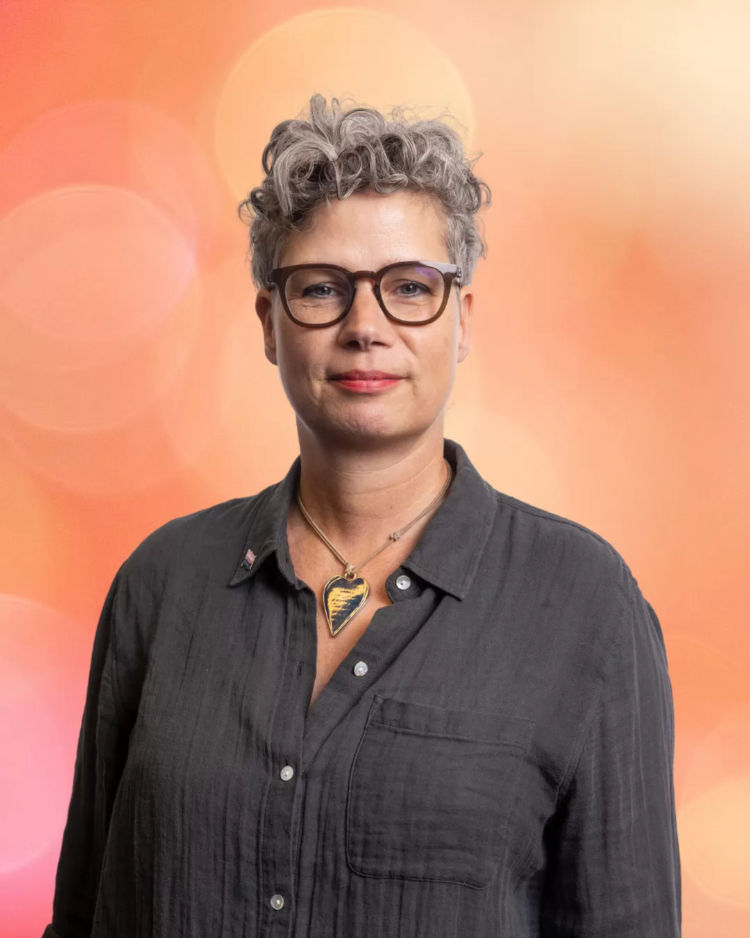 Portret van Stella. Een persoon met kort krullend grijs haar staat voor een oranje achtergrond en draagt een bruin montuur bril, een hartvormige ketting en een donkergrijze blouse.