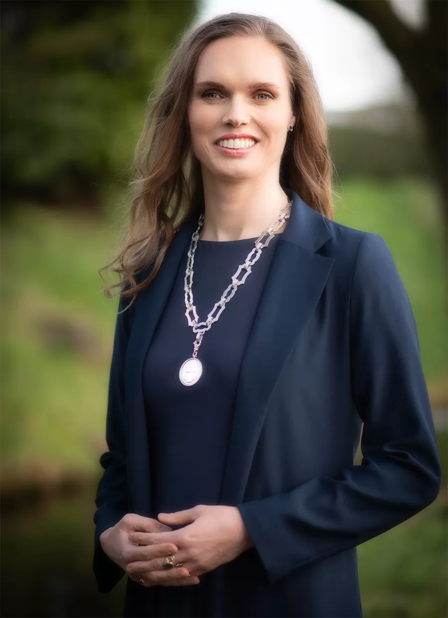 Vrouw met lang donker haar draagt een donkerblauwe blazer. Ze heeft een ambtsketen om en staat in een groene omgeving.