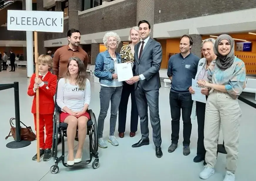 Negen mensen presenteren de petitie in de lobby van de Tweede Kamer. Een kind in de groep houdt een protestbord vast met de tekst: "Pleeback!"
