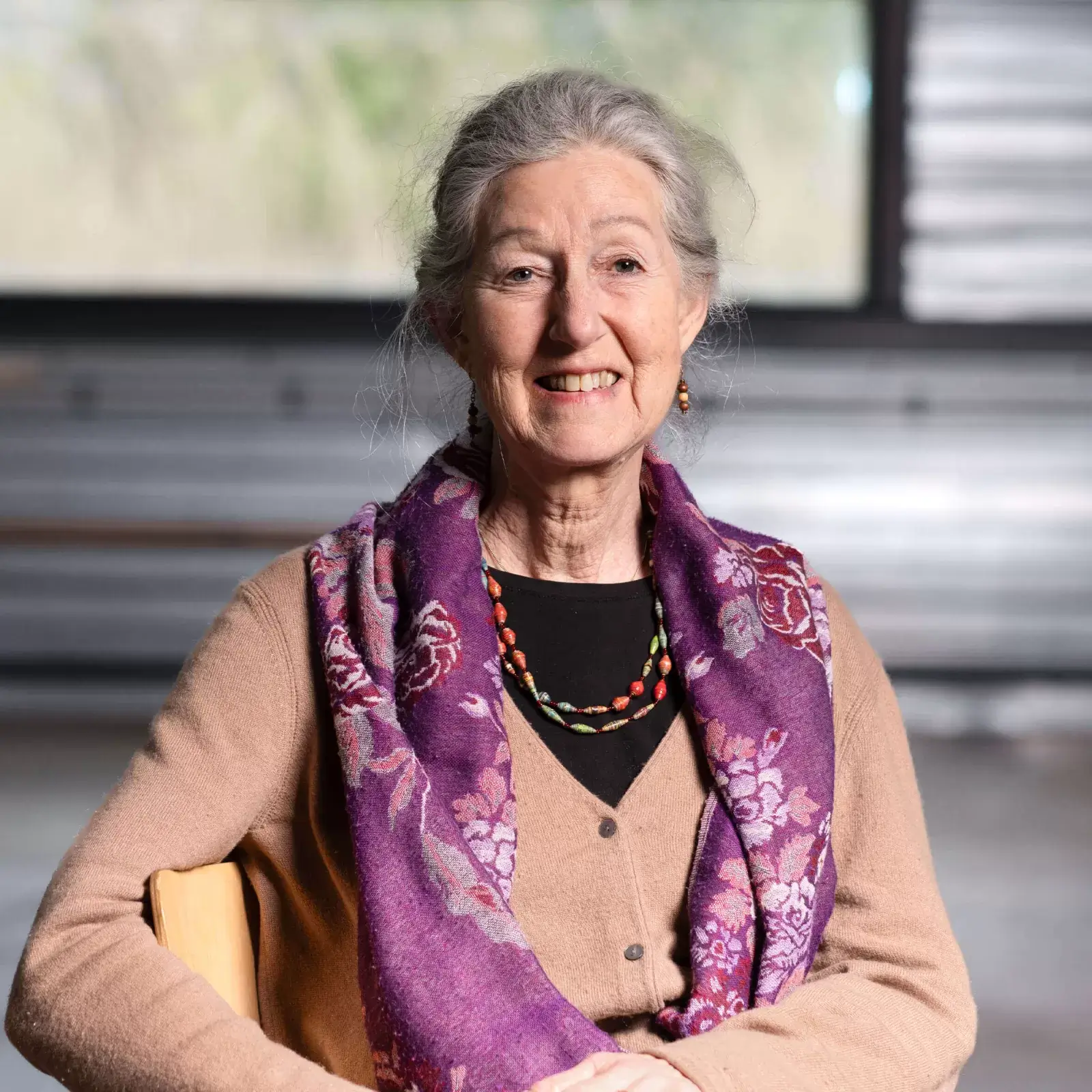 Portret van Ina Vijzelman. Een witte vrouw zit ontspannen op een stoel en kijkt je lachend aan. Ze heeft grijs haar, draagt een lichtbruin vest, een paarse sjaal en een subtiele meerkleurige kralenketting.