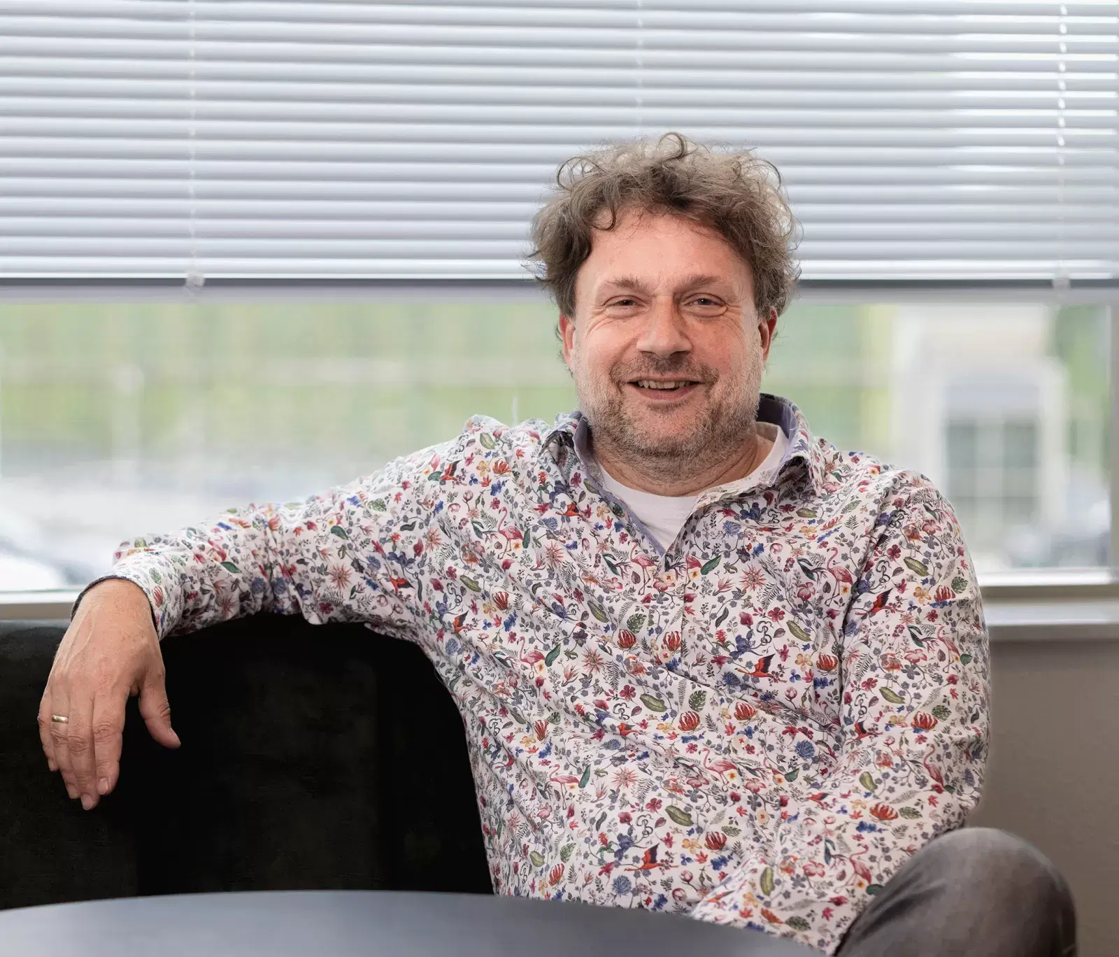 Een vrolijk kijkende man met een lichte huidskleur, krullend donkerbruin haar en een stoppelbaard. Hij draagt een meerkleurig overhemd en zit op een bank, met één arm ontspannen leunend op de rugleuning.