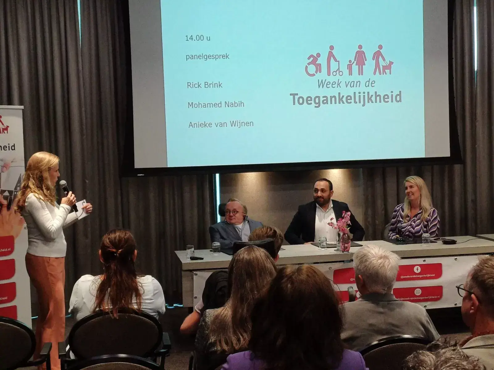 Zaaltje met publiek. Op het podium zitten drie mensen achter een tafel. Zij kijken naar de gespreksleider die naast hen op het podium staat.