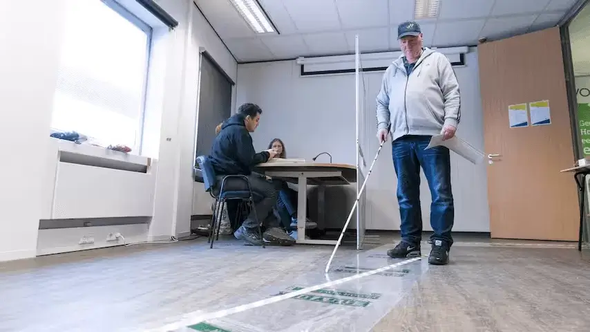 Persoon met een witte stok zoekt zijn weg in een kantoorruimte. Op de achtergrond zitten twee mensen aan een bureau te werken.