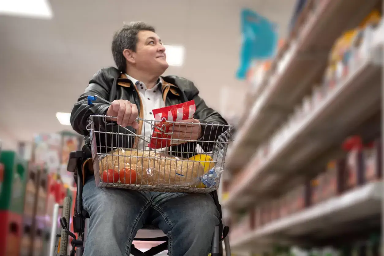 Een witte persoon in een handrolstoel in een supermarkt, draagt een zwart jack en spijkerbroek. In het mandje op schoot zitten alledaagse levensmiddelen, zoals tomaten en pasta.