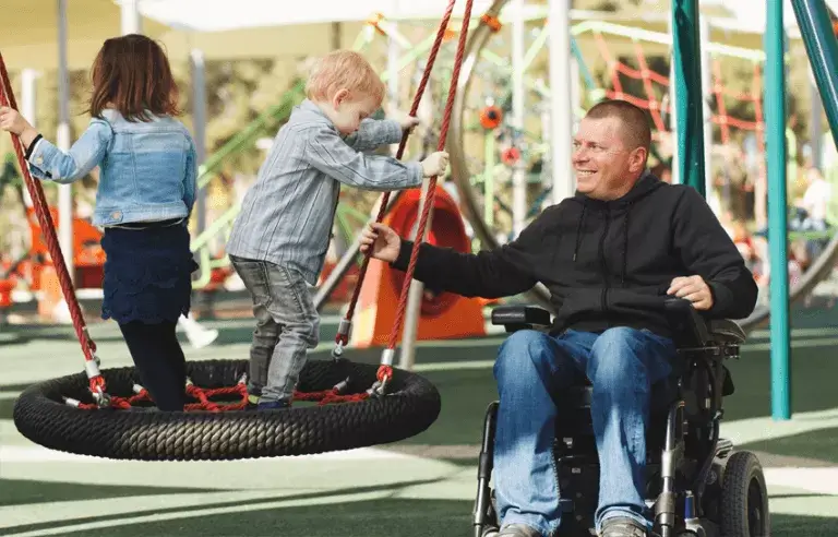 Twee kinderen staan op een nestschommel. Een man in een elektrische rolstoel duwt de schommel met één hand en kijkt vertederd. Op de achtergrond zijn meer speeltoestellen zichtbaar.