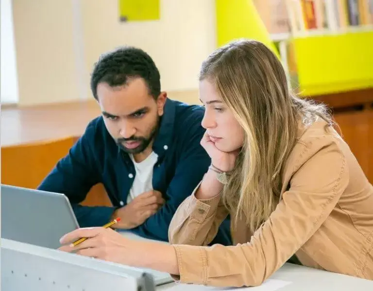 Een jonge vrouw met een lichte huidskleur en een jonge man met een licht getinte huidskleur zitten aan een bureau en kijken naar dezelfde laptop. Op de achtergrond is een boekenkast zichtbaar.