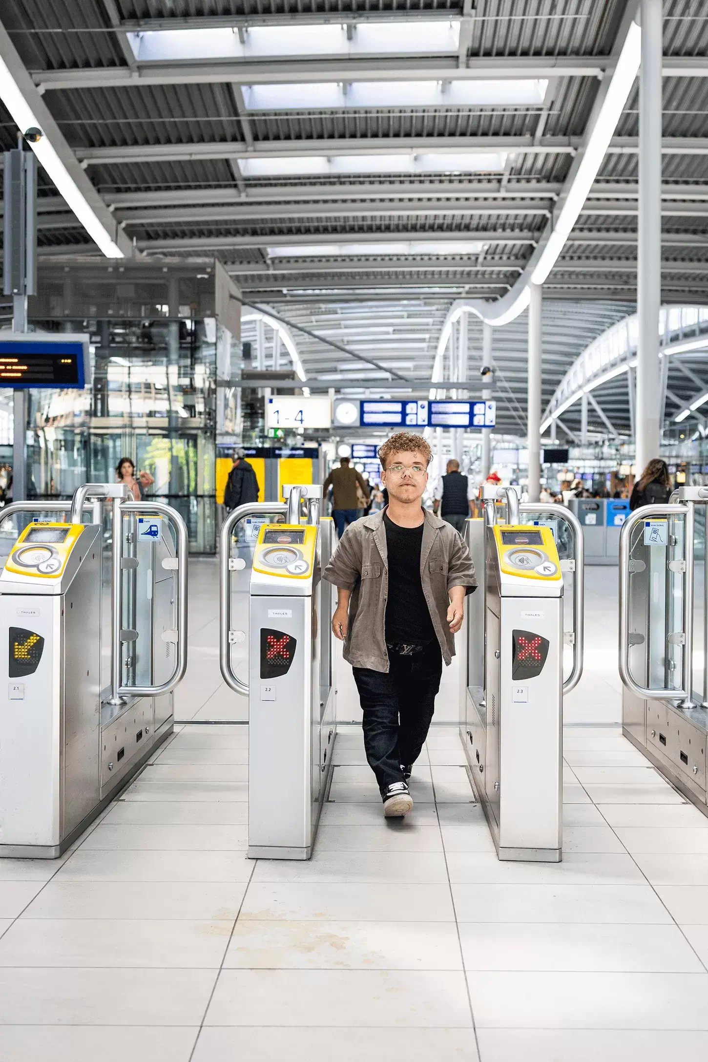 Een klein persoon met een lichte huidskleur stapt door een open toegangspoortje op station Utrecht Centraal. Hij kijkt recht in de camera. Hij heeft korte blonde krullen en draagt een bril, gympen, een donkere jeans, een zwart shirt met erover een bruin overhemd met korte mouwen. De poortjes naast hem zijn gesloten. Op de achtergrond zijn een aantal blauwwitte informatieborden en het grijze plafond van de stationshal zichtbaar.