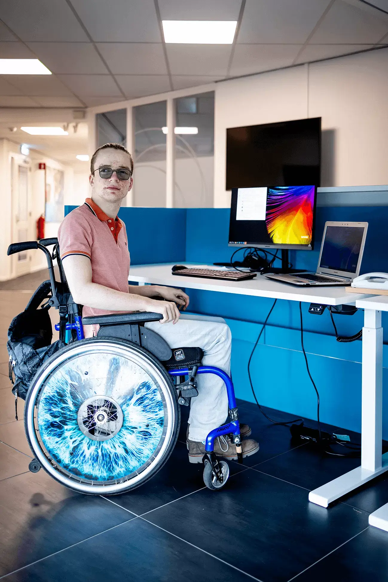 Een jonge man zit in een blauwe rolstoel aan een witte tafel met daarop een laptopscherm en toetsenbord. Zijn blik is op een punt naast de camera gericht. Het blauwe wiel van de rolstoel valt op. Op de binnenkant staat een uitvergrote foto van zijn eigen oog.  Hij heeft een lichte huid en zijn haar in een staart met opgeschoren zijkanten. Hij draagt een blauwe bril, een rood gestreept shirt en witte broek. Rond het bureau staat een blauwe scheidingswand.