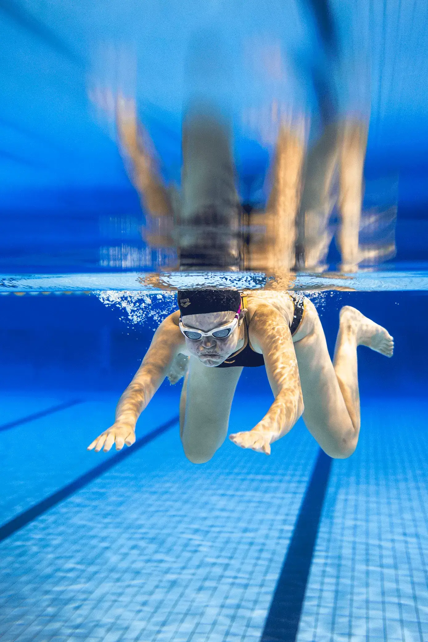 De foto is onder water genomen. Een jonge vrouw zwemt net onder het wateroppervlak. Ze heeft een lichte huidskleur en draagt een donker badpak, een witte duikbril en donkere badmuts. Het licht weerkaatst op haar huid en de bodem van het zwembad. Door het water wordt alles in de achtergrond blauw. Het wateroppervlak geeft een vage weerspiegeling van de vrouw en bodem weer.