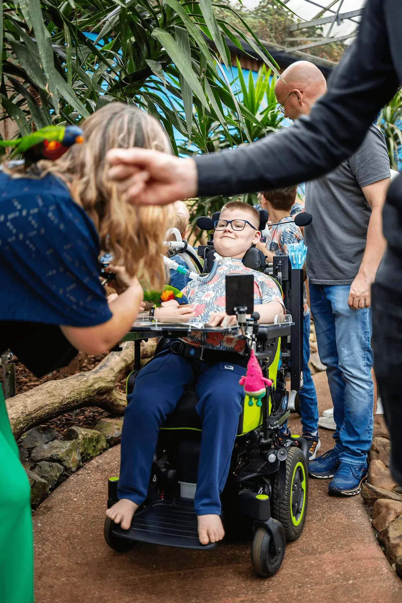 Een jongen met een lichte huidskleur in een aangepaste elektrische rolstoel staat in een binnenruimte in een dierentuin. Hij kijkt geïnteresseerd naar wat er om hem heen gebeurt, terwijl een roodgroenblauwe parkiet voer uit zijn hand eet. Voor hem staat iemand met lang blond haar en een parkiet op de schouder. Iemand reikt zijn hand uit voor de camera langs om de parkiet op diens hand te laten stappen. Achter de rolstoel staan meerdere mensen en naast hem staan grote tropische planten.