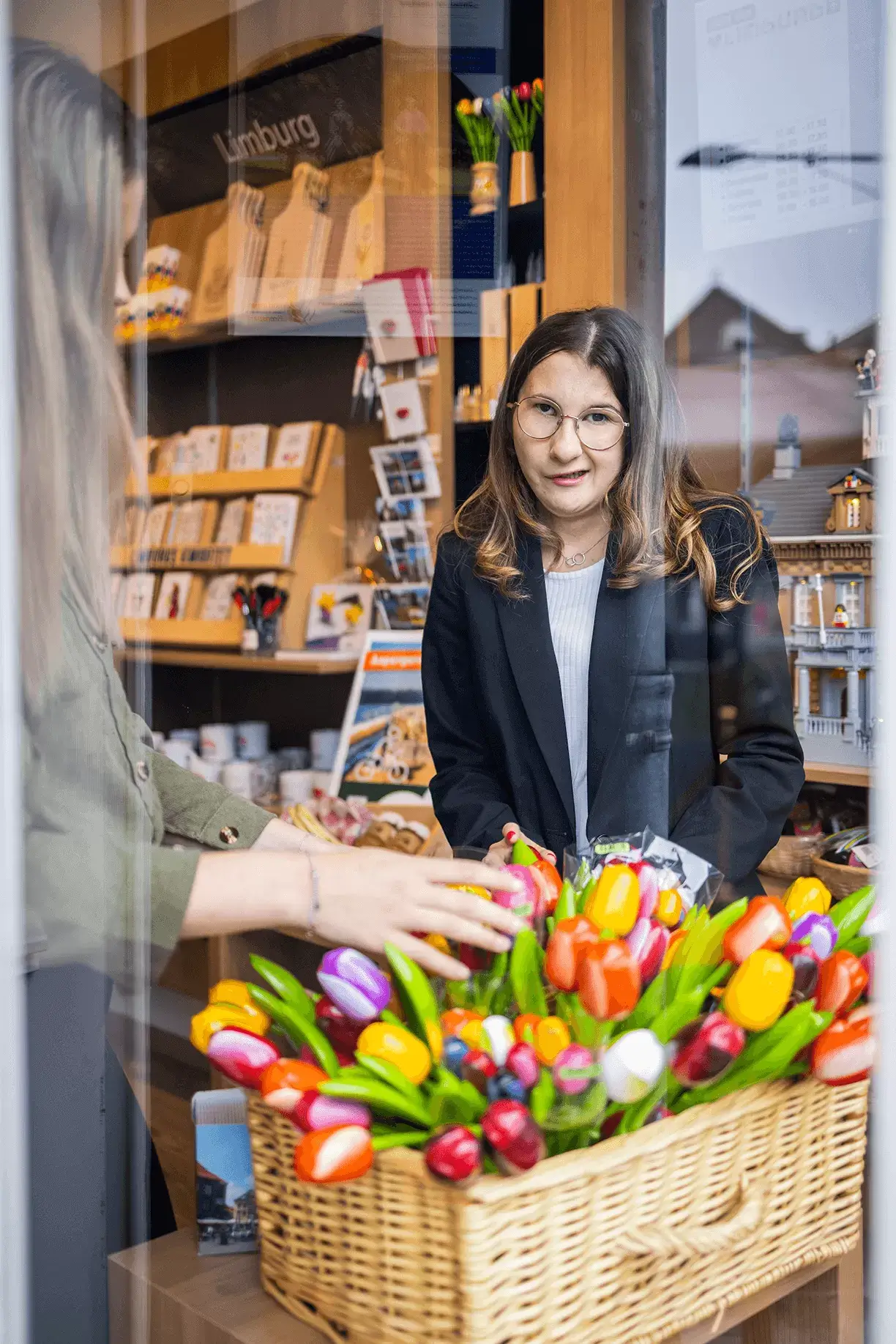 Een jonge vrouw kijkt door een raam in de camera. Een deel van haar gezicht is verlamd. Ze heeft een lichte huid en bruin lang haar. Ze draagt een zwart jasje, daaronder een wit shirt en een bril. Ze staat in de winkel van de VVV toeristeninformatie Hart van Limburg. Voor haar staat een rieten mand met kleurige tulpen uit kunststof tegen het raam. Links staat een andere vrouw met haar handen in de tulpenmand. Rechts op de foto wordt het huis aan de overkant van de straat in het raam weerspiegeld.