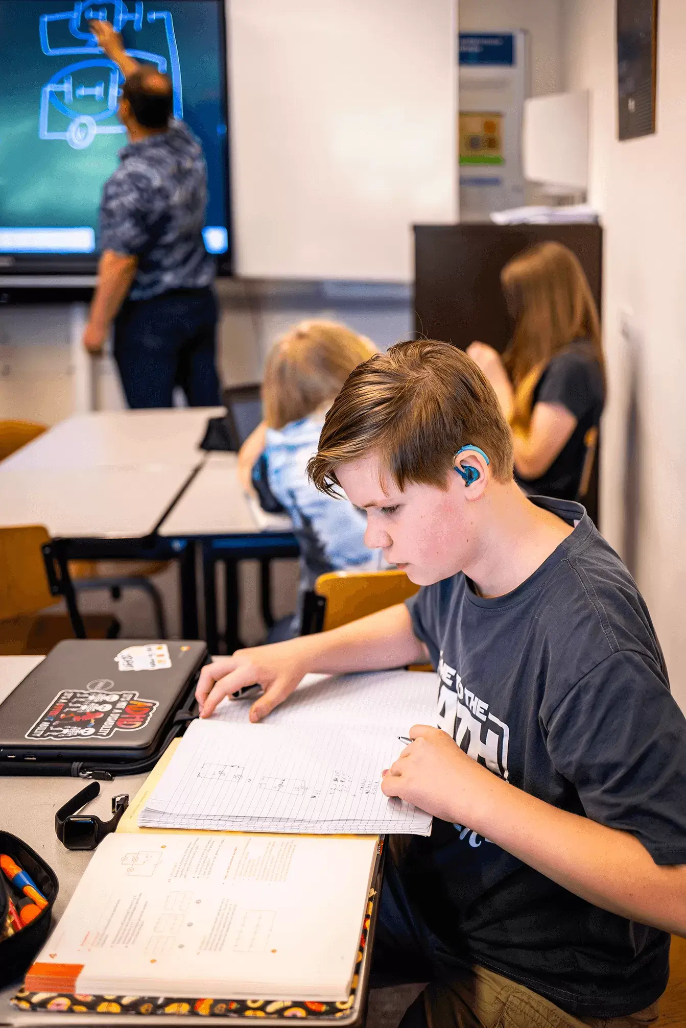 Een jongen met lichte huidskleur en kort donkerblond haar zit aan een schooltafel en leest een groot schoolboek. Hij draagt een oordopje en een donkergrijs shirt. Op de achtergrond staat een docent die iets op het bord tekent. Voor de docent zitten twee klasgenoten samen aan een schoolbank. De gezichten van de docent en klasgenoten zijn niet te zien.