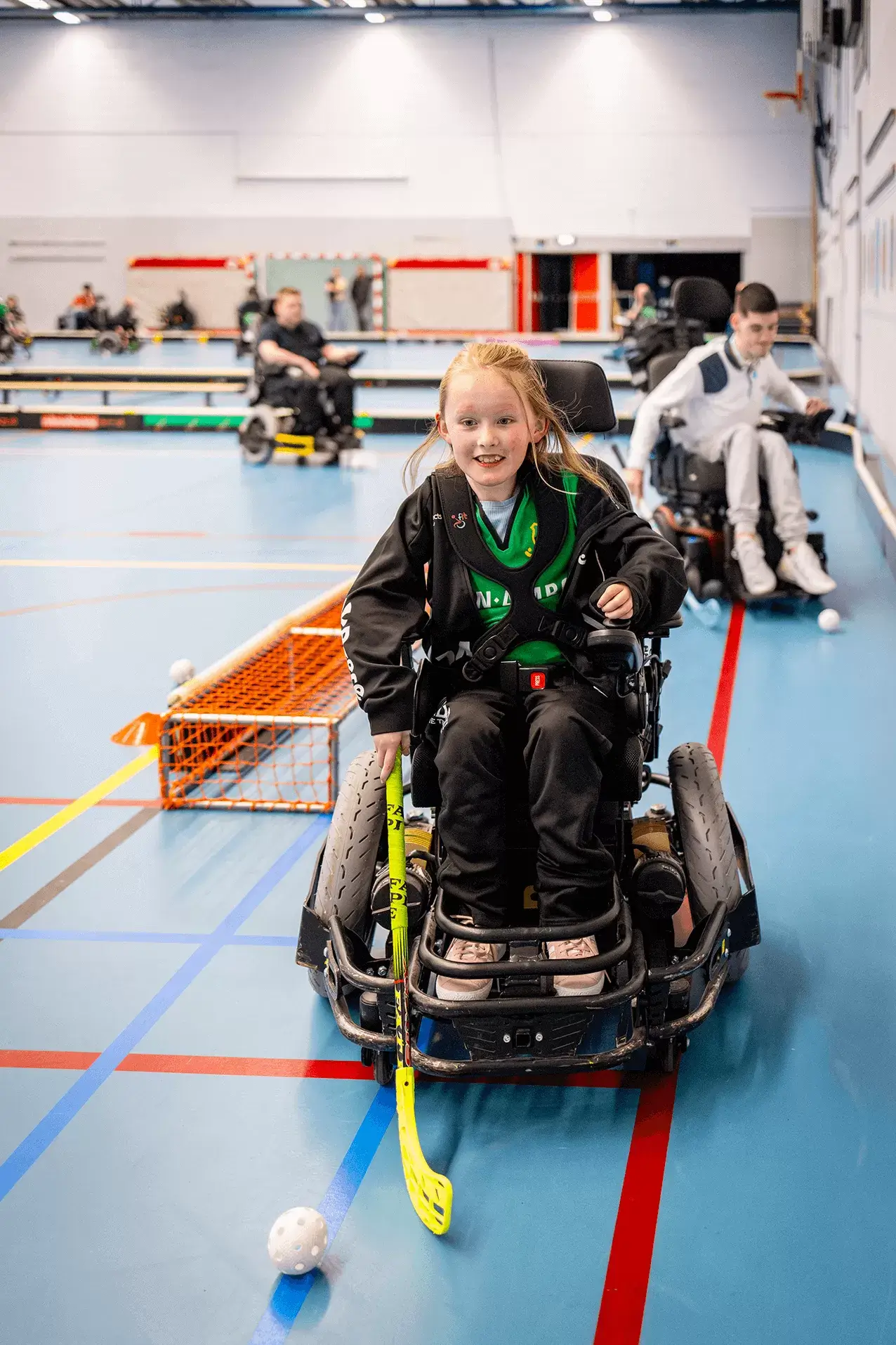 Een meisje van negen jaar zit in een elektrische rolstoel. Met haar rechterhand houdt ze een gele hockeystick vast, met haar linkerhand bestuurt ze de rolstoel. Ze lacht enthousiast en haar blik is gericht op het witte hockeyballetje voor de stick. Ze heeft een lichte huid en rood haar in een staart. Ze draagt een zwarte broek, groen shirt en zwart jasje. Op de achtergrond zijn twee andere kinderen in rolstoelen met hockeysticks te zien. Ze trainen in een sportzaal met witte muren en lichtblauwe vloer. 